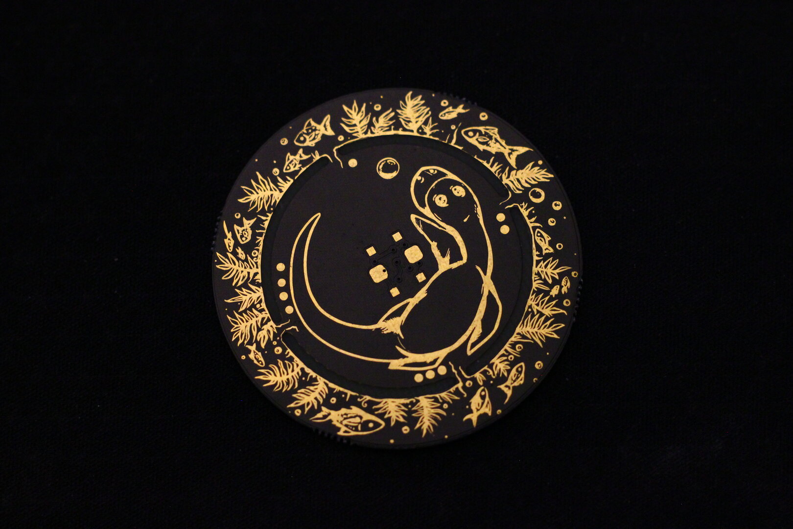 Front of the Nessie, a black circular PCB with a cartoonish Loch Ness monster in gold, surrounded by underwater flora and fauna